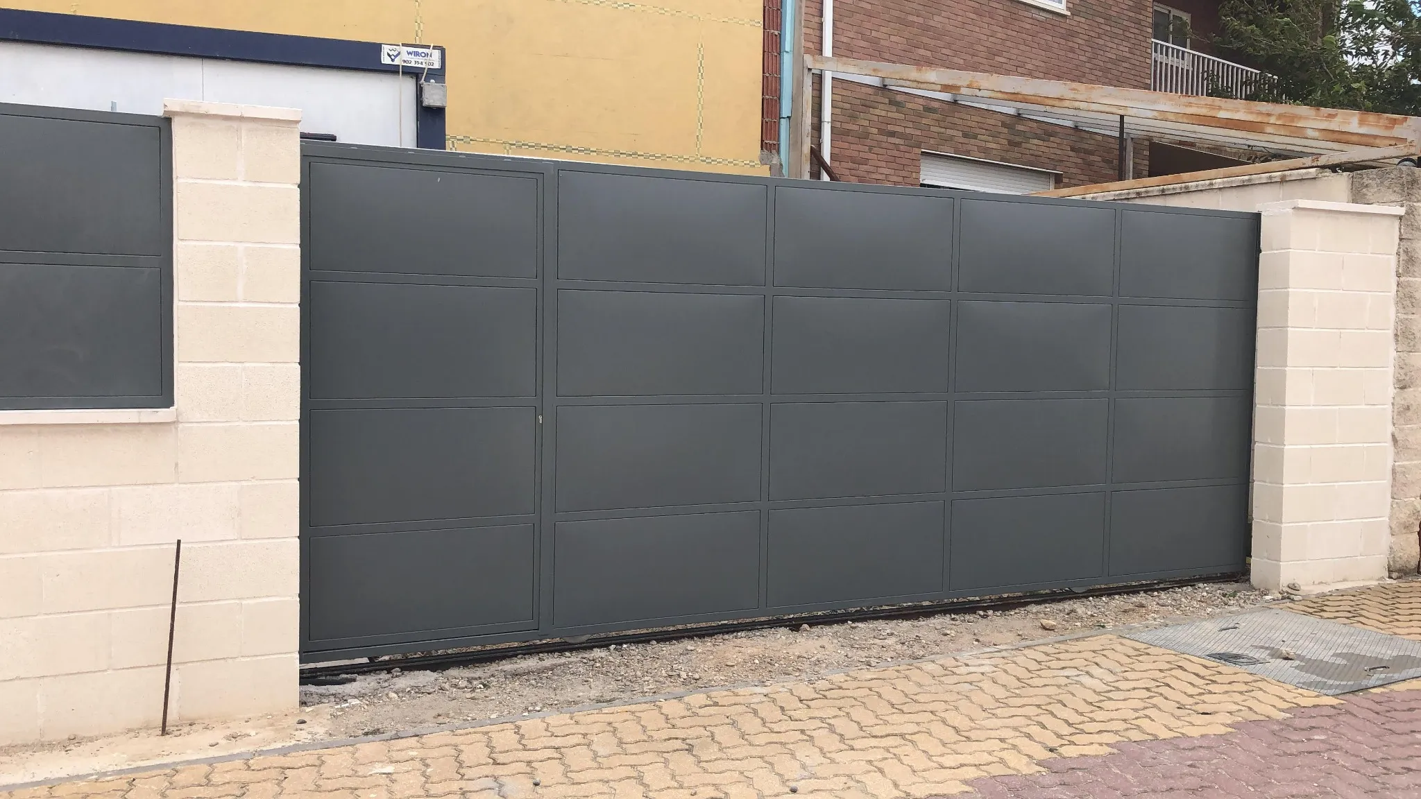 Puerta corredera metálica gris instalada entre pilares de bloque claro.