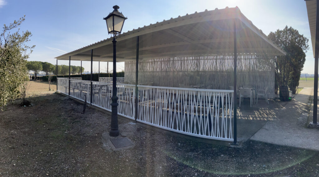 Pérgola metálica blanca con techo a dos aguas y barandillas decorativas, situada en un espacio exterior con mesas y sillas, rodeada de árboles y campo abierto.