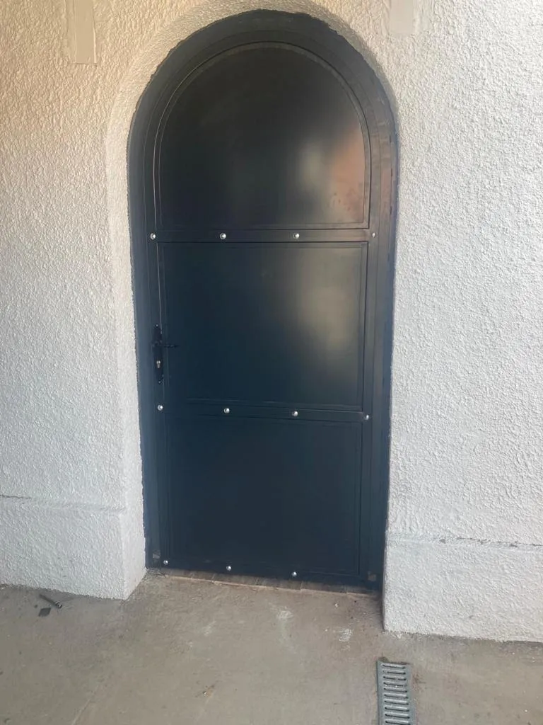 Puerta metálica con arco superior, pintada en negro, en pared blanca rugosa